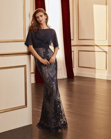 Mujer posa con un vestido largo azul oscuro decorado, en un ambiente elegante y luminoso.