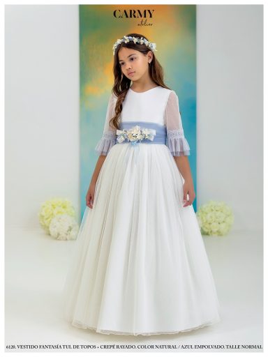 Niña con vestido blanco, mangas de tul y cinta azul en la cintura, posando elegantemente.