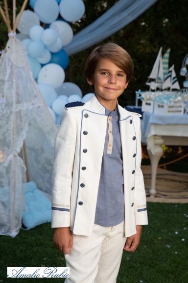 Niño vestido de traje blanco con detalles azules, sonríe en un ambiente festivo.