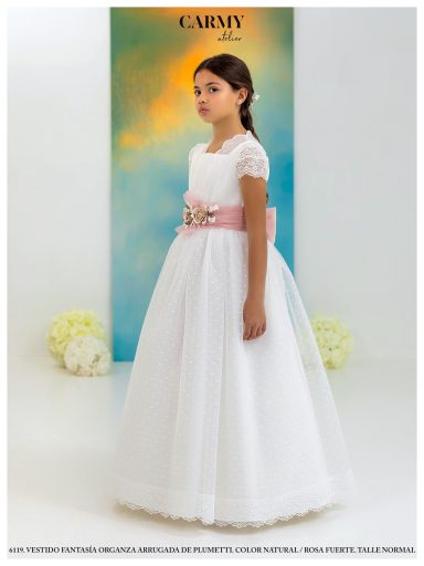 Niña luciendo un vestido blanco con cinturón rosa, de pie en un fondo azul claro.