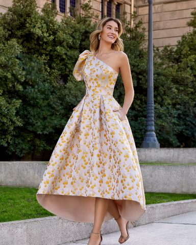 Mujer sonriente con vestido amarillo y flores, de un solo hombro, en un entorno urbano.