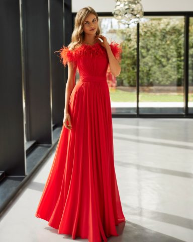 Vestido largo rojo con detalles de plumas en las mangas, de pie en un ambiente luminoso.