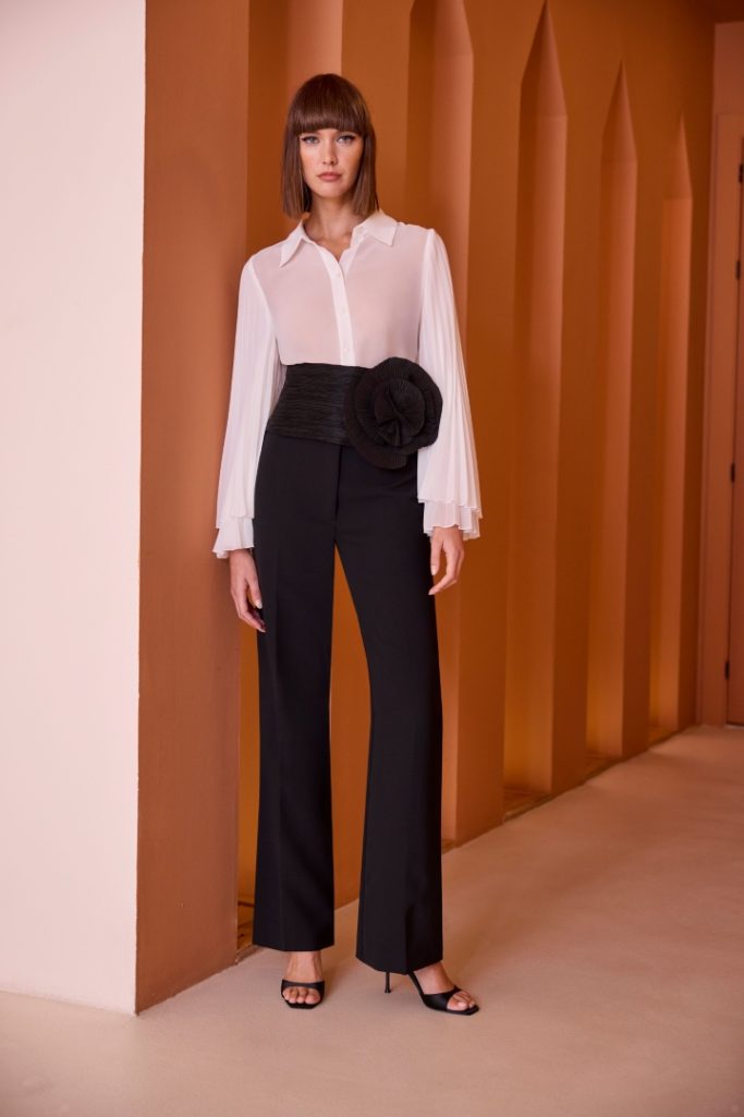 Modelo de pie con camisa blanca y pantalones negros, decoración en forma de flor.