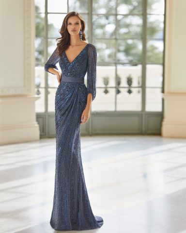 Mujer con vestido largo azul y mangas, frente a un fondo con ventanas grandes.