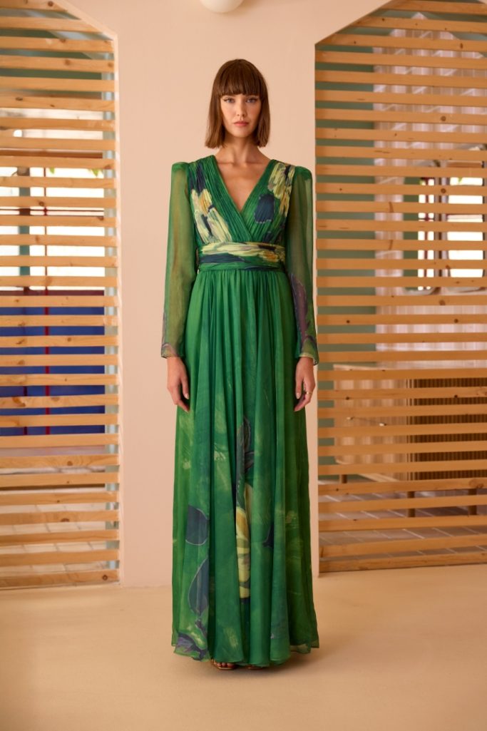 Mujer de pie con vestido largo verde y estampado, fondode madera clara.