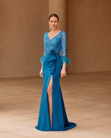 Mujer con vestido largo azul, mangas de encaje y abertura en la falda, posando en un ambiente elegante.