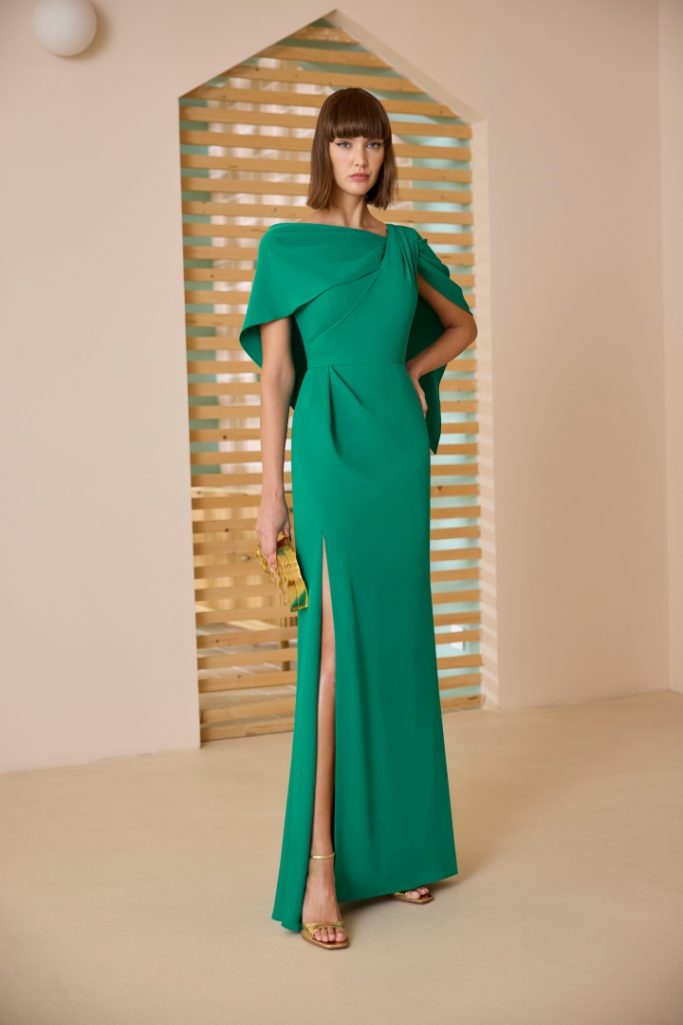 Mujer de pie con un elegante vestido verde, detalle de abertura lateral y fondo minimalista.