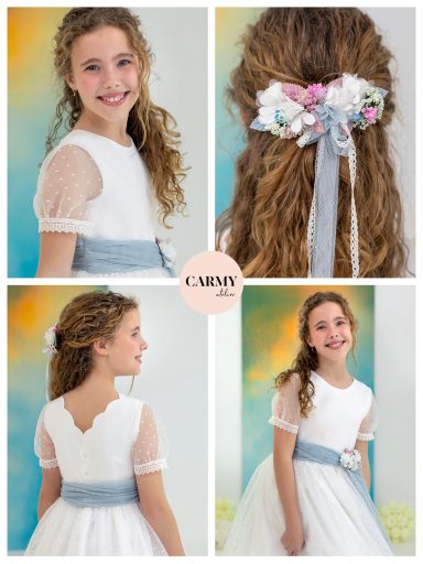 Niña sonriente en un vestido blanco con detalles en la parte superior y un lazo floral en el cabello.