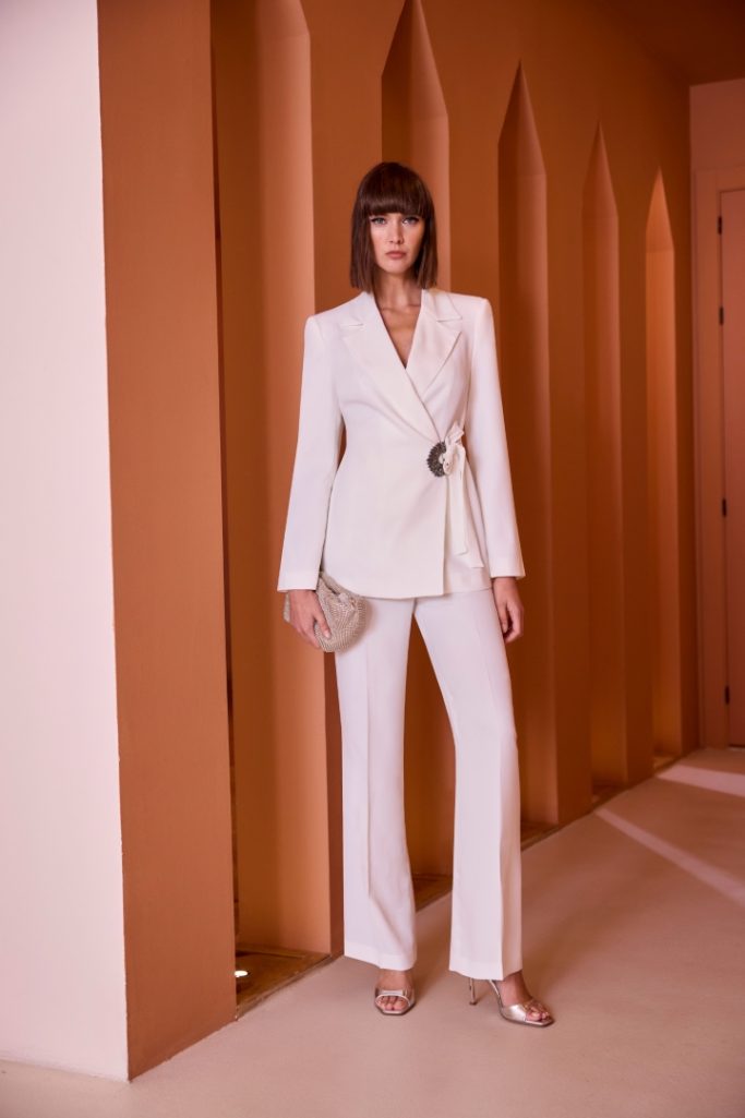 Mujer con traje blanco, bolso y zapatos elegantes, posando en un entorno de tonos cálidos.